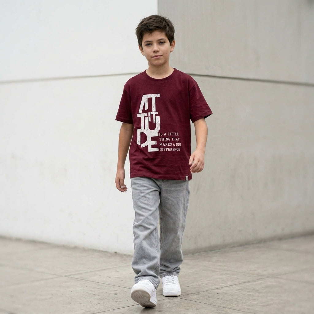 Niño con polo guinda Attitude de ANG Kidz, estampado blanco frontal y manos en los bolsillos del jean gris.